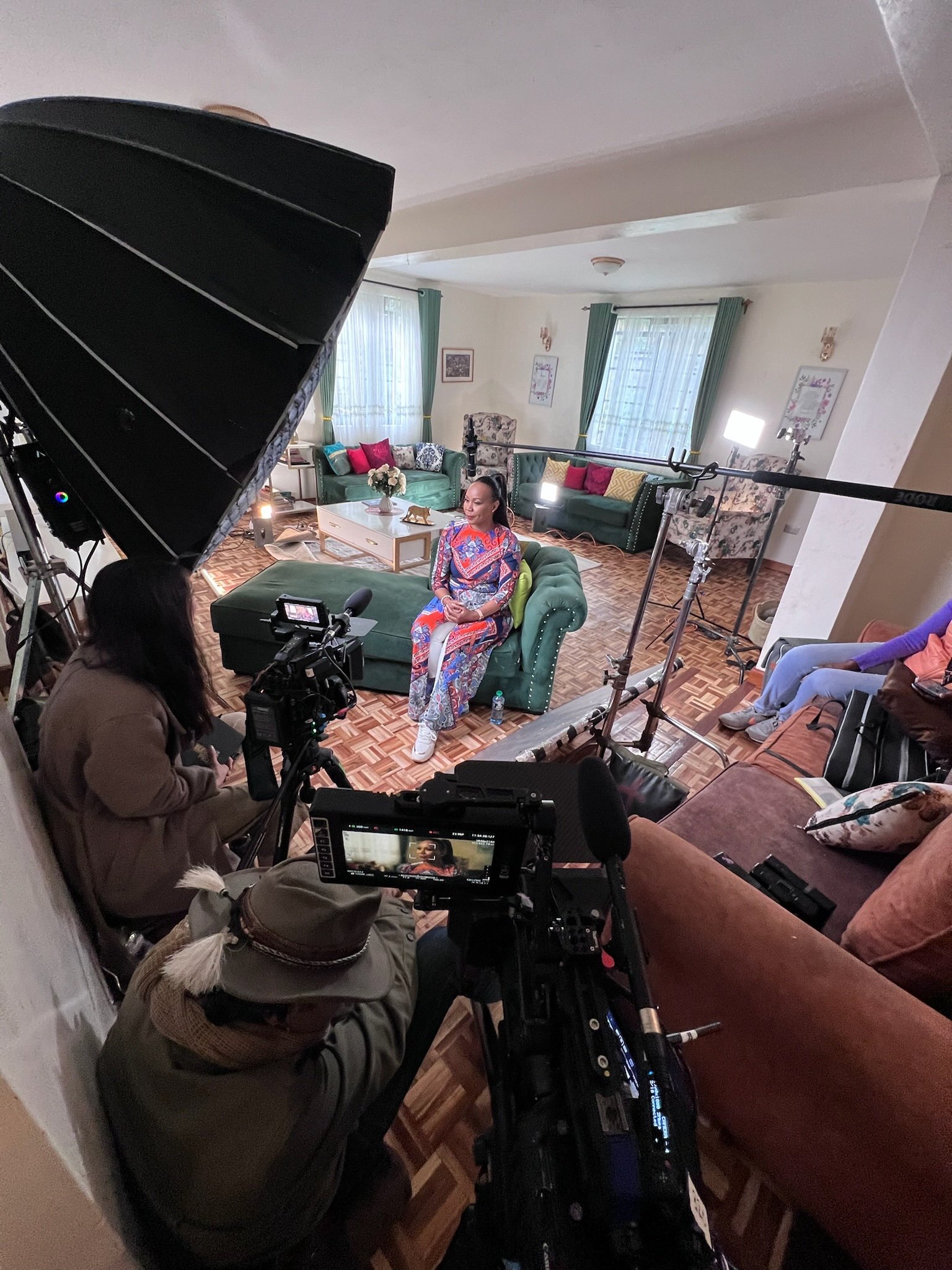 A film crew records an interview in a well-lit living room. A woman in a red and blue dress sits on a green couch, smiling as she speaks. Around her, the crew operates cameras, lighting, and a boom mic, while screens and equipment are visible on a nearby table. The setting includes bookshelves, warm lighting, and a painting of an archway on the wall.